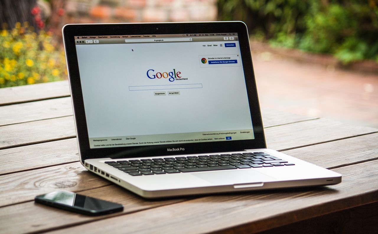 Un MacBook Pro affichant Google Search sur une table en bois à l’extérieur, à côté d’un smartphone.